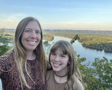 A woman and a girl smile for a selfie, standing in front of a scenic river vista with lush green trees and hills under a clear blue sky.