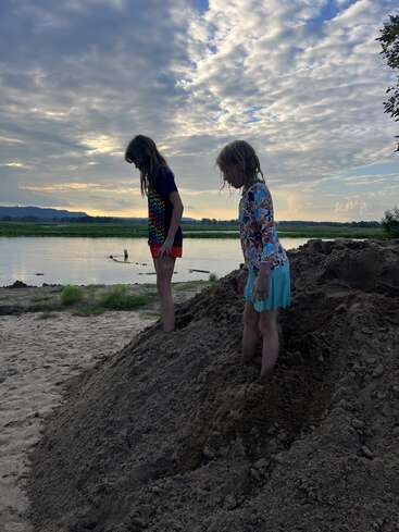 Two children stand on a sandy mound near a river at sunset. Cloudy sky above, peaceful scenery, and playful summer atmosphere. Nature and innocence blend beautifully.