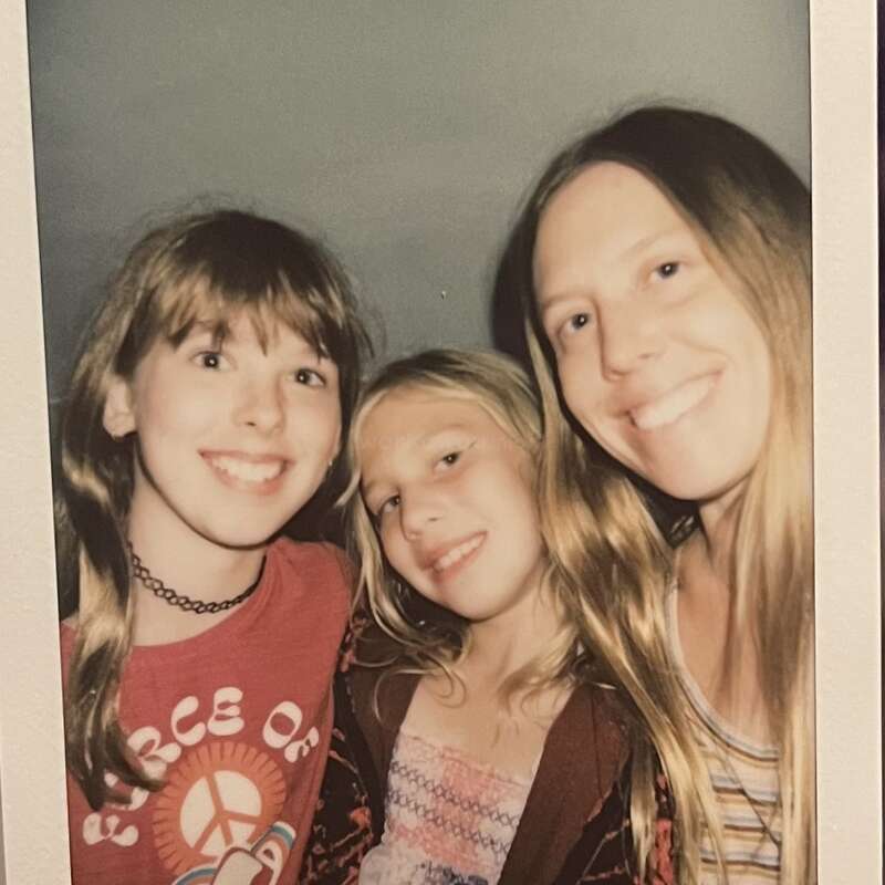 Three smiling girls with long hair pose closely together for a photo. They look happy and relaxed. Bright clothing and friendly expressions highlight their joyful moment.