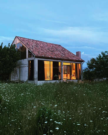 Uma casa aconchegante com telhado de telhas fica em um campo de flores silvestres ao entardecer. Luzes quentes brilham por dentro, criando uma atmosfera acolhedora e tranquila.