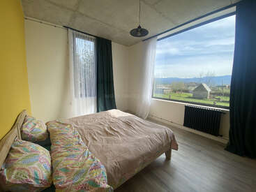 Ein gemütliches Schlafzimmer mit einem großen Fenster bietet einen tollen Blick auf die ländliche Umgebung. Es gibt ein Doppelbett mit gemusterten Kissen, Holzböden und eine minimalistische, moderne Einrichtung.