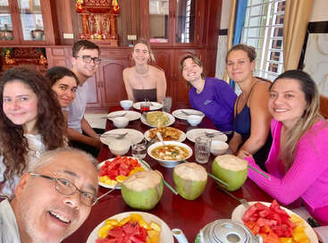 Siete personas se sientan alrededor de una mesa de comedor, sonriendo para una selfie, disfrutando de una comida con frutas tropicales, cocos y varios platos deliciosos en una acogedora habitación.