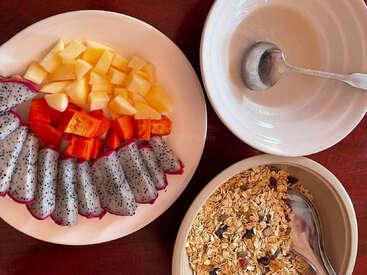 Un desayuno saludable compuesto por rodajas de pitaya, papaya y manzana en un plato, muesli seco en un bol y un bol vacío con una cuchara.