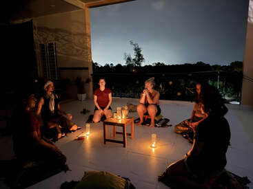 Seis personas se sientan en círculo en un patio por la noche, iluminadas por velas en el suelo y la mesa, creando un ambiente acogedor, íntimo y meditativo.