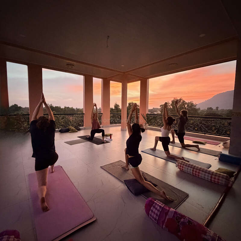 Seis personas practican yoga sobre colchonetas en un espacio abierto cubierto al atardecer, rodeadas de naturaleza, montañas y cálidos cielos anaranjados, creando una atmósfera tranquila.