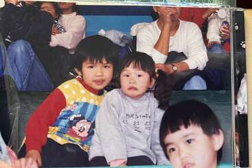 Duas crianças sentadas juntas em primeiro plano, uma sorrindo e a outra parecendo insegura. Outras pessoas, incluindo crianças e adultos, estão em segundo plano assistindo a algo.