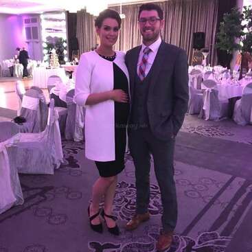 Un couple bien habillé pose ensemble lors d'un événement élégant. La salle est décorée de tables rondes, de chaises blanches et d'un éclairage violet, créant une atmosphère festive.