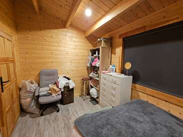Cette chambre douillette en bois comprend un fauteuil, un lit, une commode, des étagères remplies de vêtements, un tabouret et une grande fenêtre avec des stores.