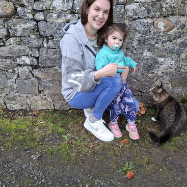 Eine Frau und ein kleines Mädchen mit einem Schnuller posieren an einer Steinmauer im Freien und kauern neben einer flauschigen Katze auf grünem Gras und Kiesboden.