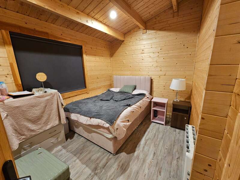 Cette chambre douillette en bois comprend un lit simple avec une couverture grise, une tête de lit rose, des tables de chevet, une lampe, une commode couverte et une fenêtre recouverte d'un store.