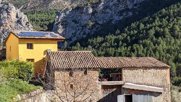 A rustic house with yellow walls and solar panels sits beside an old stone building, nestled in a lush green forested mountain landscape under bright sunlight.