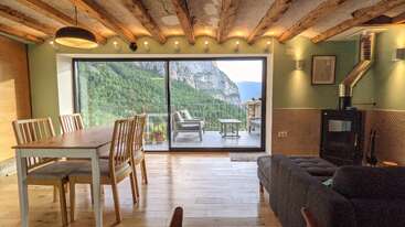 This cozy living room features wooden beams, modern furniture, and a wood stove. Large glass doors open to a balcony with breathtaking mountain and forest views.
