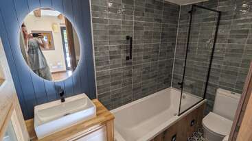 This modern bathroom features a bathtub with a glass partition, green subway tiles, a wall mirror reflecting a person, a white sink, wooden vanity, and toilet.
