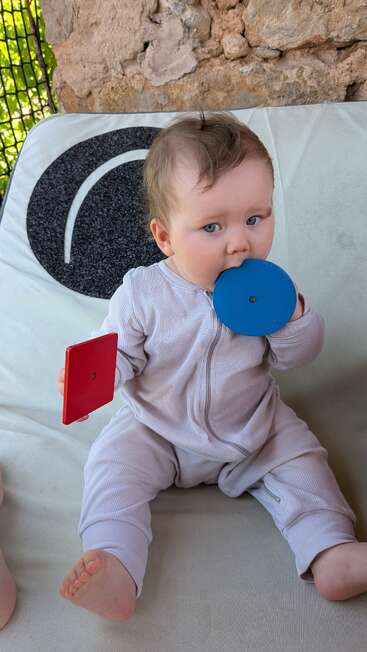 A baby with blue eyes sits on a couch, wearing a light onesie, holding a red square and chewing on a blue round object, looking curious.