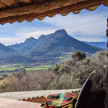 Um terraço coberto e aconchegante tem vista para uma paisagem montanhosa deslumbrante, com campos verdes ondulados, céu azul e um cobertor colorido estendido sobre uma cadeira em primeiro plano.