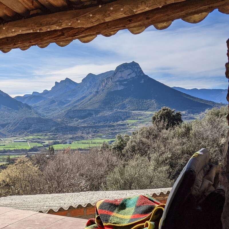 A cozy covered terrace overlooks a stunning mountain landscape with rolling green fields, clear skies, and a colorful blanket draped over a chair in the foreground.
