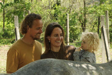 A smiling man, woman, and a young child with curly hair stand outdoors beside a gray horse, surrounded by lush green trees and rustic wooden fences.