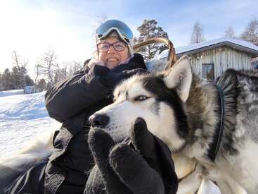Uma mulher vestindo roupas de inverno e óculos de proteção sorri enquanto abraça um cão husky. Eles estão ao ar livre em uma paisagem nevada, com uma cabana e árvores ao fundo.