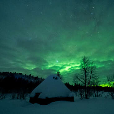 Une cabane enneigée repose paisiblement sous une aurore boréale verte et lumineuse. Des arbres dénudés et un paysage hivernal entourent le bâtiment sous un ciel étoilé.