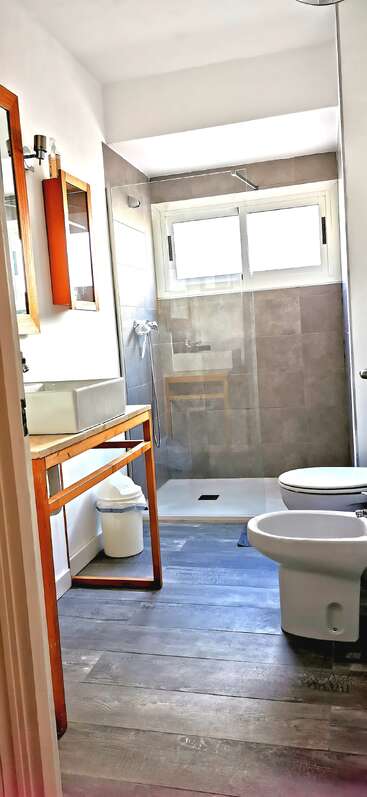 A bright, modern bathroom features a shower with glass partition, toilet, bidet, wooden vanity, vessel sink, cabinet, waste bin, and a wooden floor.