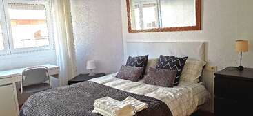 This bright bedroom features a double bed with patterned pillows, a dark throw, bedside tables with lamps, a large mirror, a desk, and folded towels.