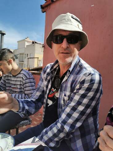 A man wearing sunglasses, a beige bucket hat, and a checkered shirt sits outdoors. He is accompanied by others, enjoying a sunny day and food.