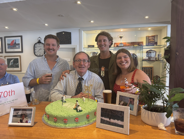 A group celebrates a 70th birthday. Three younger adults stand with an older man behind a green cake, drinks in hand, surrounded by photos and decorations.