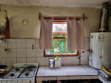 Una acogedora cocina con encimera de azulejos, cocina de gas, cafetera, ventana con cortinas, calentador de agua y vegetación exterior visible a través de la ventana, que crea un ambiente hogareño.