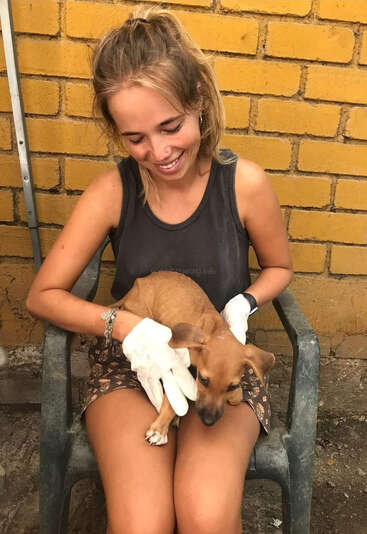 A young woman sits in a chair, cradling a small brown dog, wearing white gloves and a black tank top, with a yellow brick wall behind her.