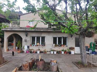 Una casa rústica de dos plantas con plantas en macetas, un árbol grande, un patio, tocones de madera, una manguera verde, un arco, ventanas y un gato negro paseando por fuera.