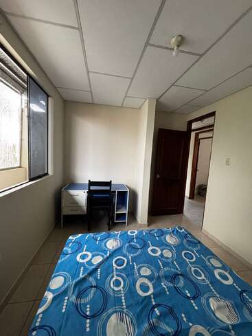 Cette image montre une chambre simple et bien rangée, avec un lit à motifs bleus, un bureau et une chaise près de la fenêtre, ainsi qu'une porte ouverte.