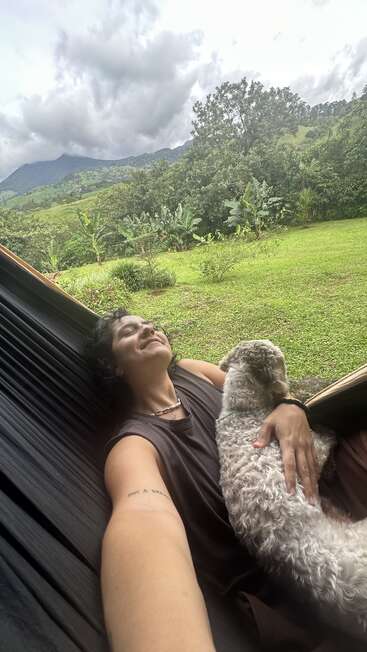 A person relaxes on a hammock, smiling and cuddling a fluffy dog. Behind them, a lush green landscape, trees, and distant mountains under a cloudy sky.