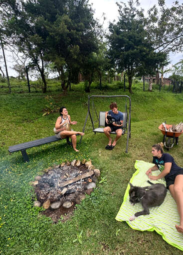 Three friends relax outdoors near a fire pit. One plays guitar, another drums, while the third lounges on a blanket beside a gray dog. Trees surround them.