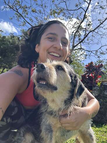 Una mujer de pelo oscuro sonríe alegremente mientras abraza a un perro desaliñado y feliz en el exterior. El cielo azul, los árboles verdes y las plantas de colores vivos crean una alegre escena al aire libre.