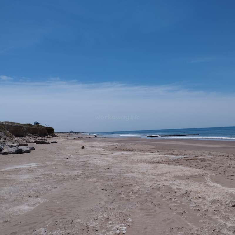 Ein ruhiger Sandstrand erstreckt sich unter einem riesigen blauen Himmel bis in die Ferne. Sanfte Wellen schlagen an den Strand, und weit entfernt sind ein paar Leute zu sehen. Eine friedliche Szene.