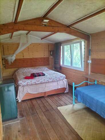 Dieses gemütliche Schlafzimmer in einer Holzhütte verfügt über ein Doppelbett, ein Einzelbett, ein großes Fenster, Holzbalken, eine kleine Kommode, eine schlichte Einrichtung und viel Tageslicht.