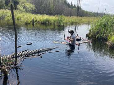 A person wearing sunglasses and gloves is sitting in a pond or swamp, working with pipes and sticks. Tall grass and trees surround the water.