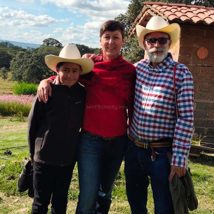 Três pessoas posam ao ar livre: um menino e um homem usando chapéus de caubói e uma mulher de vermelho. Eles estão sorrindo juntos, com um cenário rural e céu azul.
