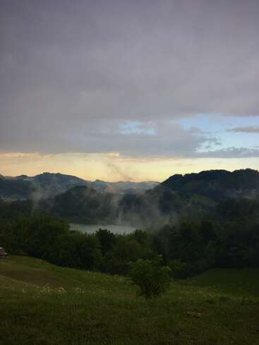 L'image représente un paysage serein composé d'un lac, d'arbres et de montagnes sous un ciel nuageux, avec une brume subtile s'élevant de la surface de l'eau.