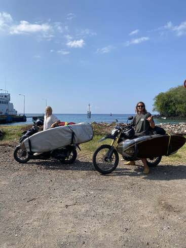 Duas pessoas com pranchas de surfe amarradas às motos posam perto da costa num dia ensolarado. Uma balsa e o céu azul compõem um cenário pitoresco.