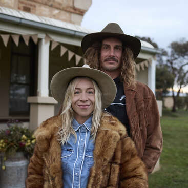 Um casal sorridente está ao ar livre, ambos usando chapéus de abas largas e roupas vintage no estilo western. A mulher usa um casaco de pele, e o homem, uma jaqueta marrom com franjas.