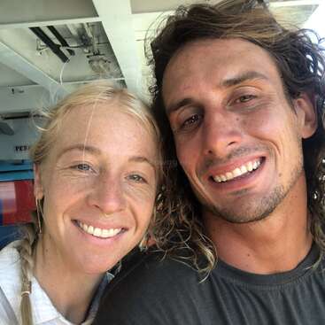 Uma mulher sorridente de cabelos loiros e um homem de cabelos castanhos cacheados tiram uma selfie em close-up juntos, parecendo felizes e relaxados num ambiente bem iluminado.