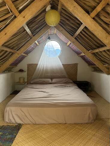 Un acogedor dormitorio abuhardillado con techo de paja, cama grande, ropa de cama de color beige suave, mosquitera, ventana redonda, farol de papel, suelo de estera tejida y decoración minimalista.