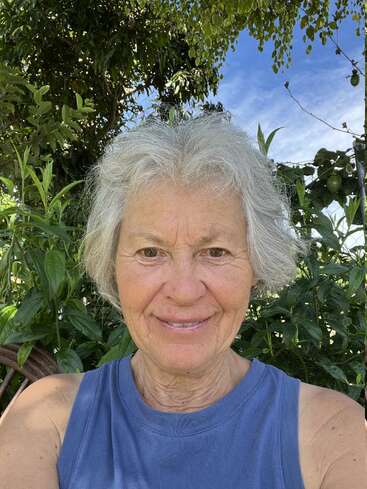 Una sonriente mujer mayor con el pelo canoso se toma un selfie al aire libre, rodeada de exuberante follaje verde y bajo un cielo azul despejado, disfrutando de la tranquilidad de la naturaleza.