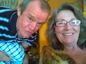 Uma mulher sorridente com óculos e um homem com uma camisa listrada posam juntos dentro de casa. Um cachorro marrom está sentado no colo da mulher, olhando para ela.