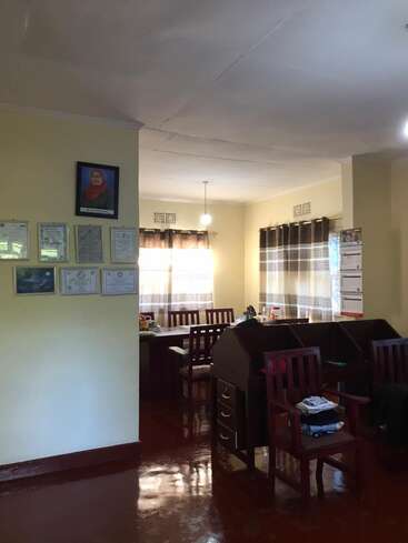 L'image représente une pièce bien éclairée avec une table à manger, des chaises et un bureau, avec une collection de certificats encadrés sur le mur et un calendrier sur le côté droit.