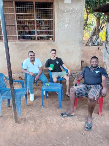 L'image représente trois hommes assis sur des chaises en plastique à l'extérieur d'un bâtiment, l'un d'eux tenant un gobelet vert, sur fond de terre et d'arbres.