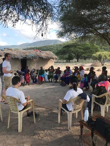 L'image représente un groupe de personnes rassemblées en cercle à l'extérieur d'une hutte, avec un homme debout s'adressant au groupe, sur fond de paysage montagneux.