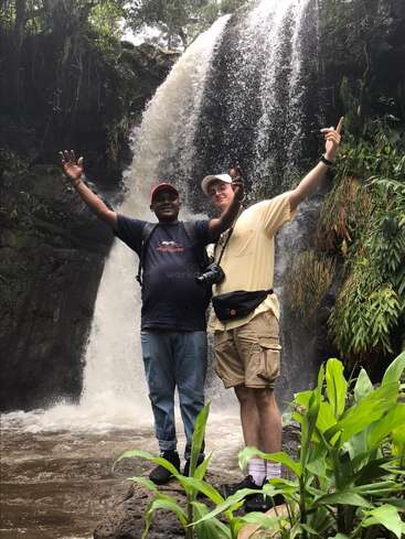 Deux hommes se tiennent devant une cascade, les bras levés, vêtus de vêtements décontractés et coiffés d'un chapeau. Ils sont entourés d'une végétation luxuriante dans un cadre naturel serein.