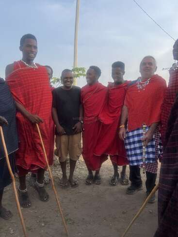 Un groupe d'hommes, dont certains portent des vêtements traditionnels masaï et des parures en perles, se tiennent ensemble à l'extérieur, souriant et prenant la pose pour une photo par une journée ensoleillée.